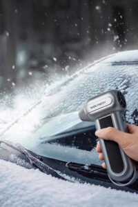 Hand holding a WOLFBOX MF100 electric air duster defroster clearing ice and snow from a car windshield during a winter snowstorm, portable cordless heater for fast windshield defogging and de-icing.